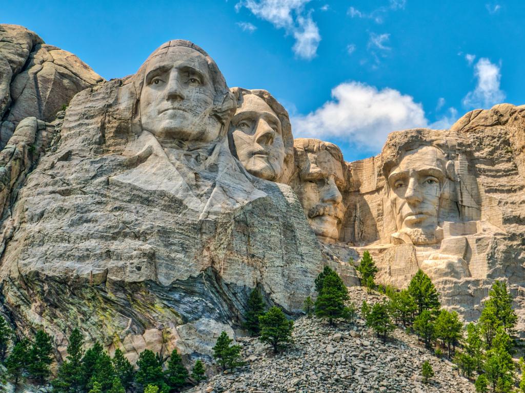 Mount Rushmore, USA with the faces of former US presidents carved into the Black Hills, trees in the foreground on a sunny day.