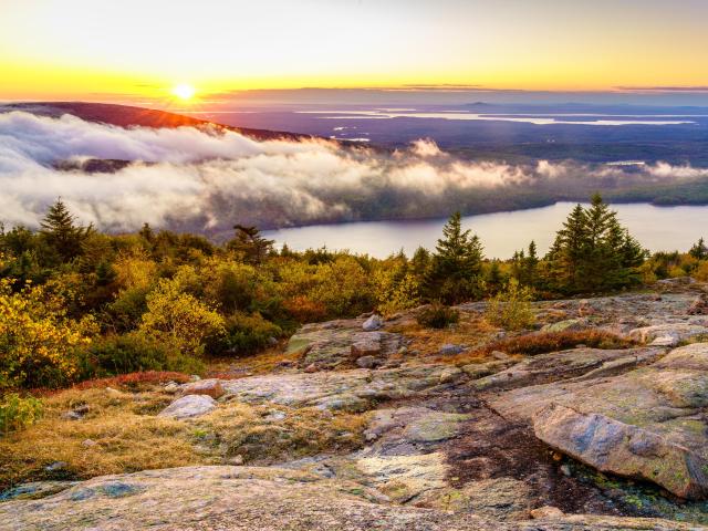 Acadia National Park, US at sunset, taken on top of a mountain looking beyond to the distance a large lake surrounded by tall trees and low cloud. 