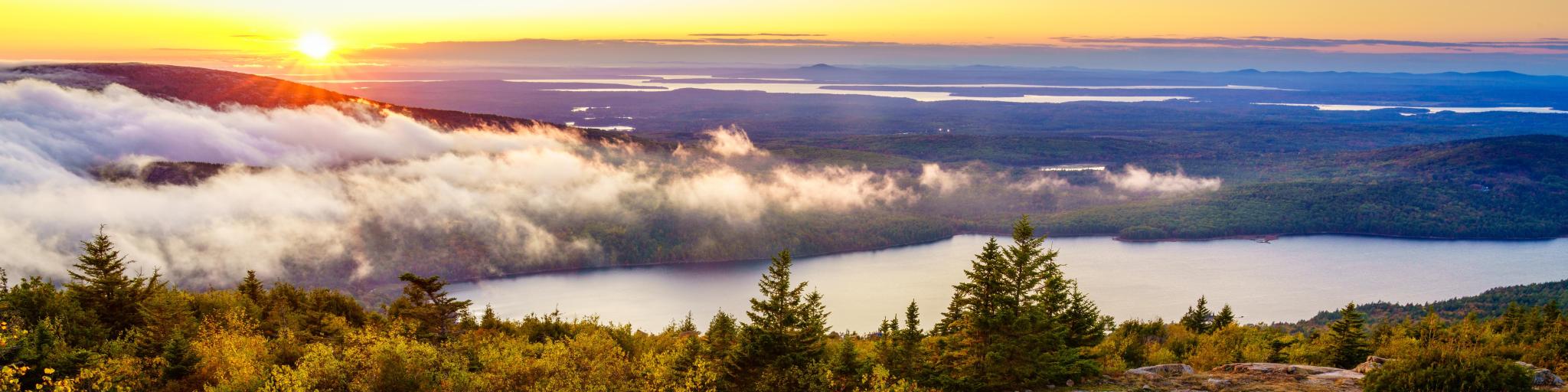 Acadia National Park, US at sunset, taken on top of a mountain looking beyond to the distance a large lake surrounded by tall trees and low cloud. 