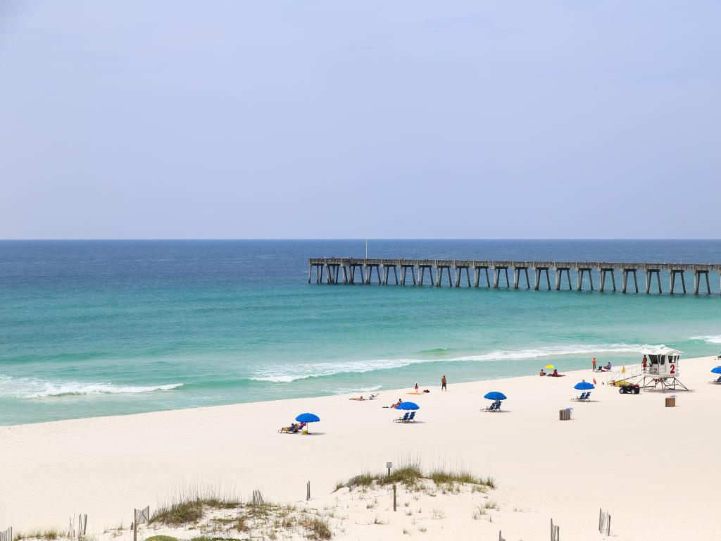 People relaxing and sunbathing on the beach with beach chairs and sunshades. In the back the Pensacola Beach Gulf Pier.