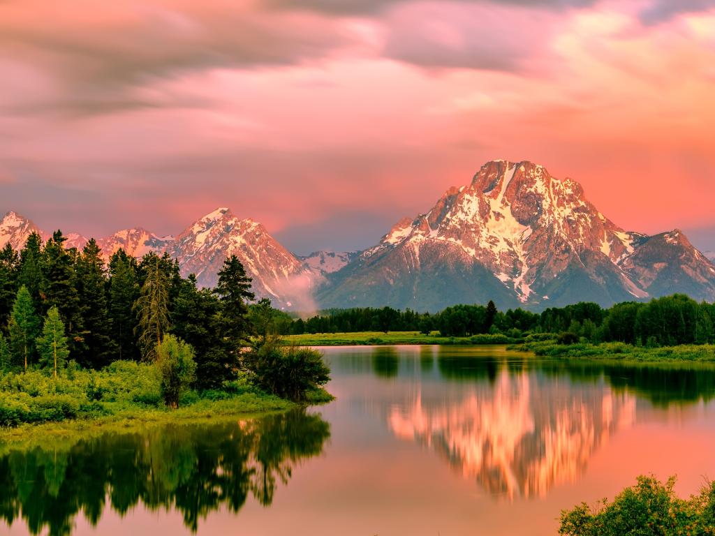 Grand Teton National Park, Wyoming with the Grand Teton Mountains in the background and the Snake River in the foreground lined with trees above a glowing red sunrise. 