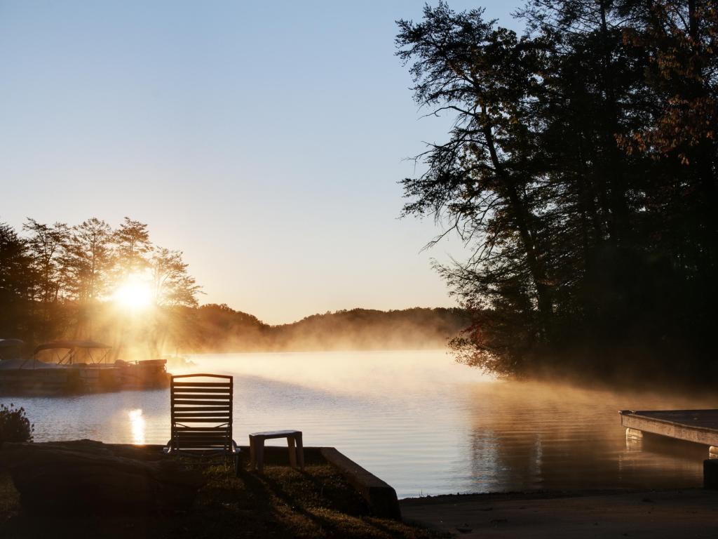 Lake Lure, North Carolina, USA with the sun rising over the lake and fog in the distance, tall trees in shadows.