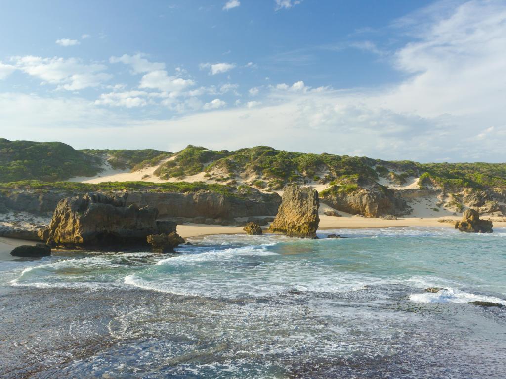 Kenton-on-Sea, South Africa taken at a rugged coast with sand and grass dunes in the distance and the sea in the foreground on a sunny day.