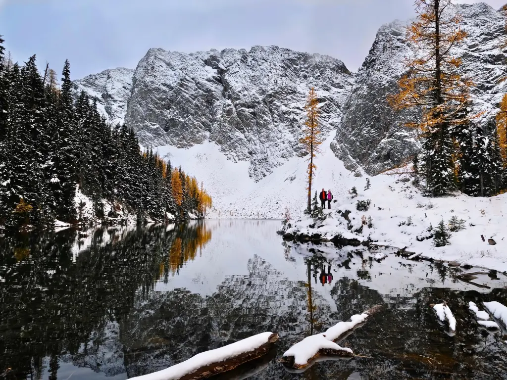 People hiking in North Cascades National Park. Tranquil lake with reflections of peaks and trees covered with snow. 