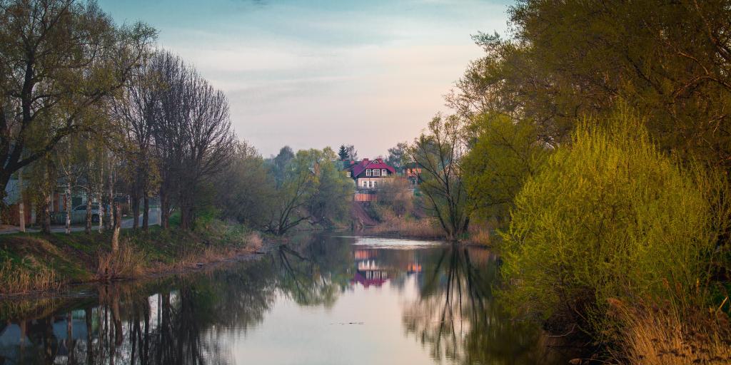 A house in the distance with its reflection visible in the water below it, surrounded by trees, in Suzdal, Russia