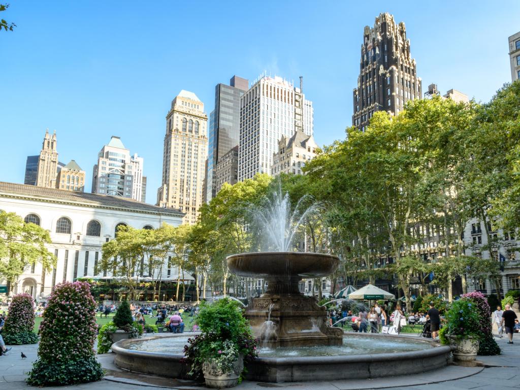 Close up of the fountain, with parkland in the background and people strolling and relaxing in Bryant Park, New York