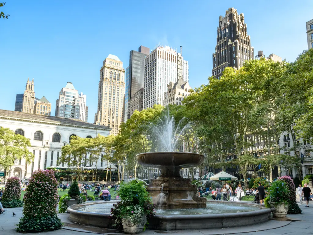 Close up of the fountain, with parkland in the background and people strolling and relaxing in Bryant Park, New York