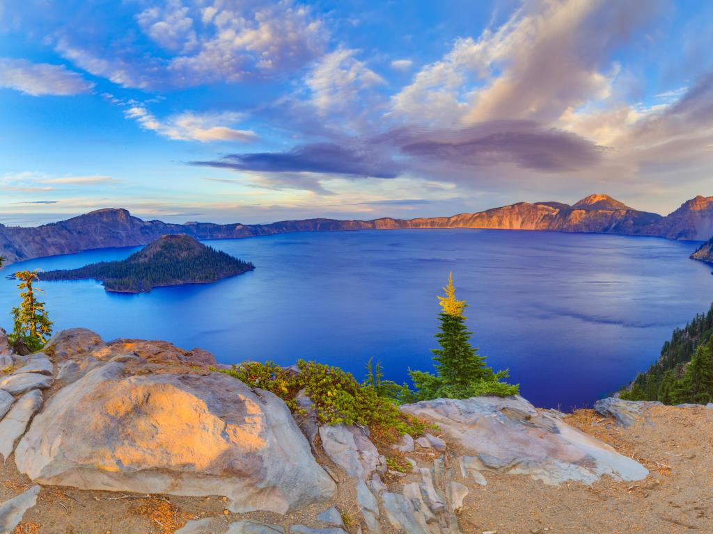 Crater Lake National Park, Oregon, USA taken at early sunset with rocks in the foreground and the lake and mountains in the distance with a blue sky above.
