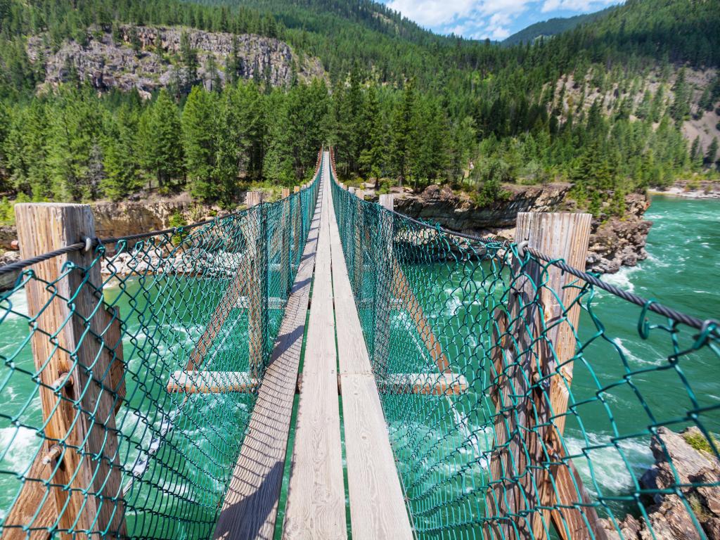 View looking over the hanging Suspension Bridge at Kootenai Falls, with lush greenery on the other side of the riverbank