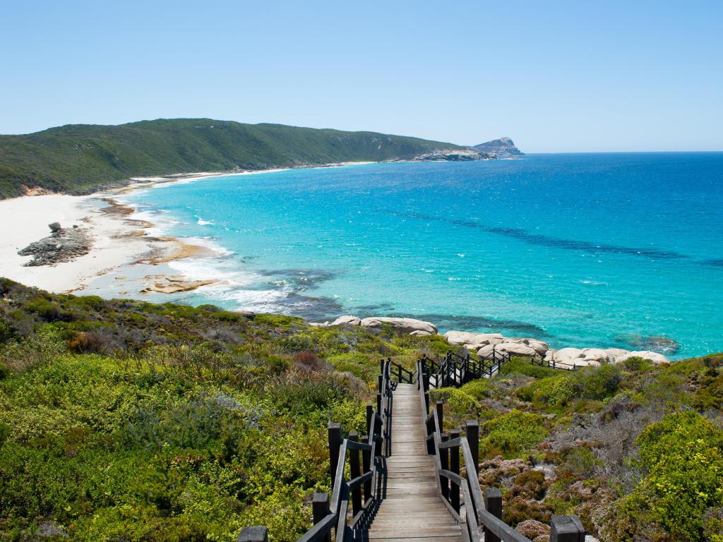 A walkway leading to a white sand beach and bright blue waters surrounded by mountains in the distance