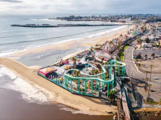 Aerial drone shot of Santa Cruz Beach Boardwalk, with the colourful amusement park next to the beach with mountains in the background.