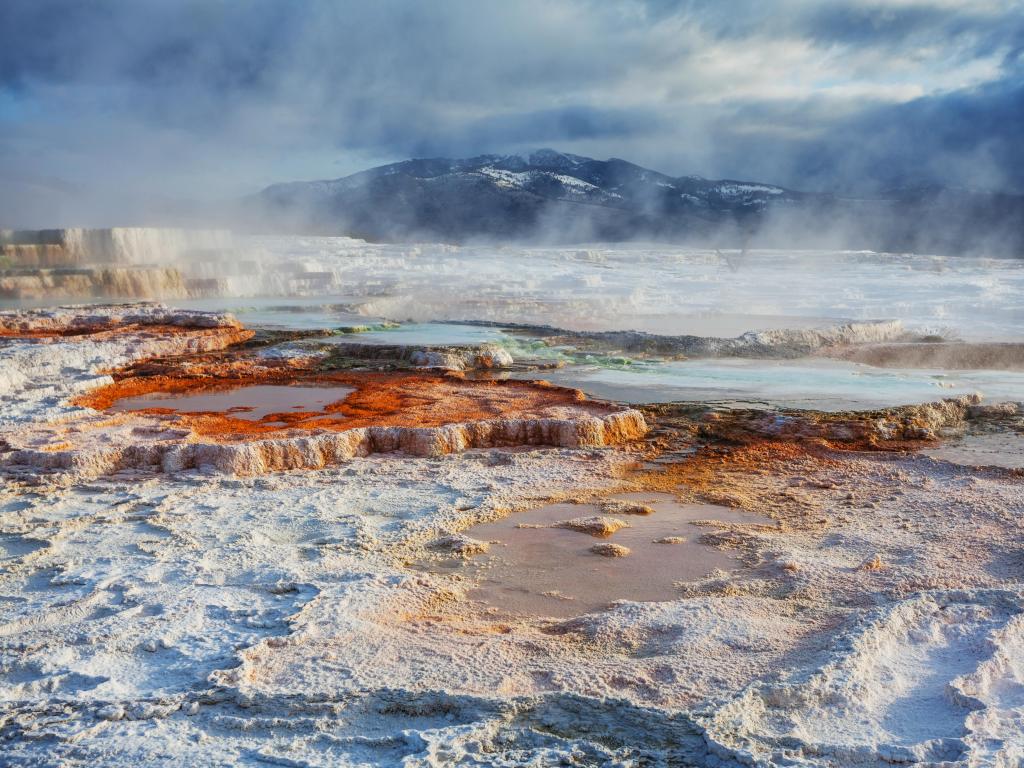 Steam coming off choppy water with vibrant rust coloured ground covered in light snow, with mountains behind