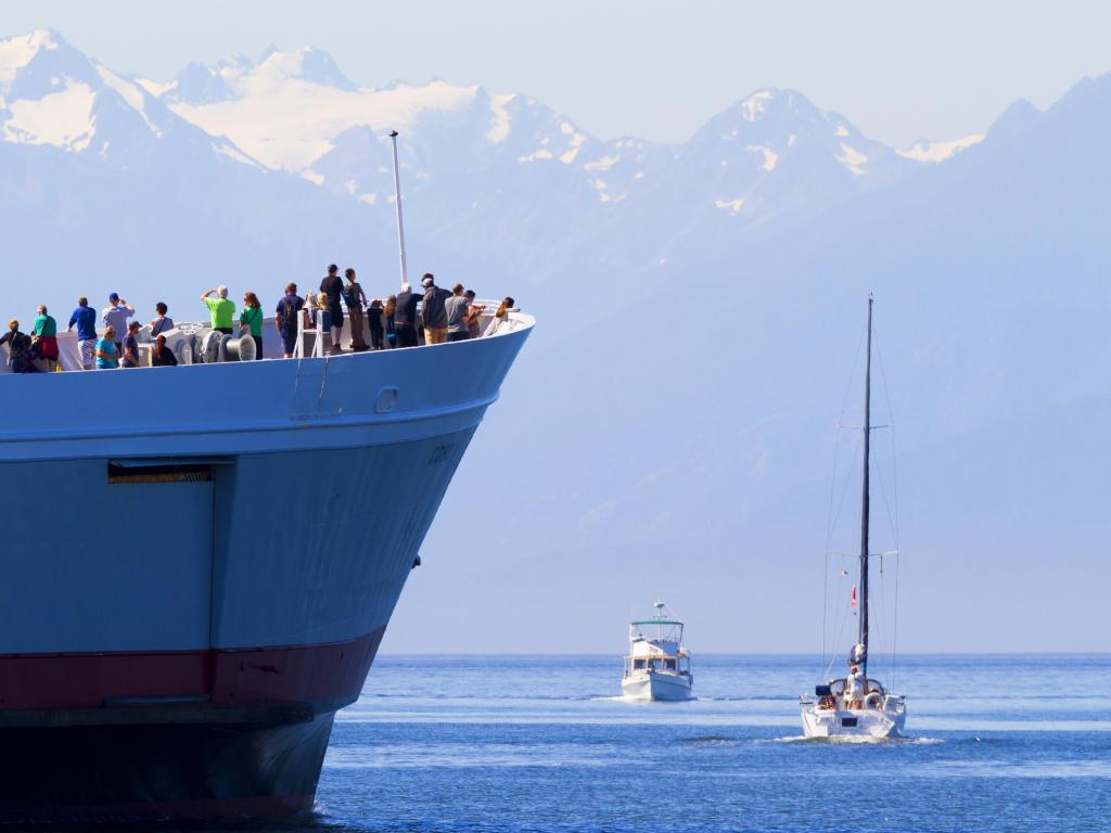 An image of people standing on a ferry with two smaller boats in the water with snowy mountains in the background