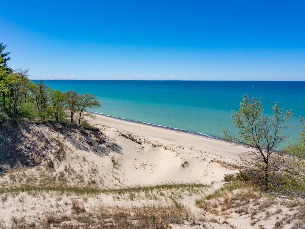 Sand dune leading down to bright blue sea with blue sky and a few trees