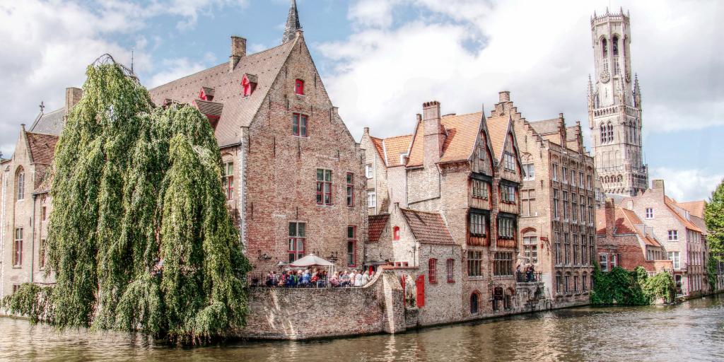 People stand under an umbrella at a restaurant on the river in Bruges, Belgium, with the belfry in the distance