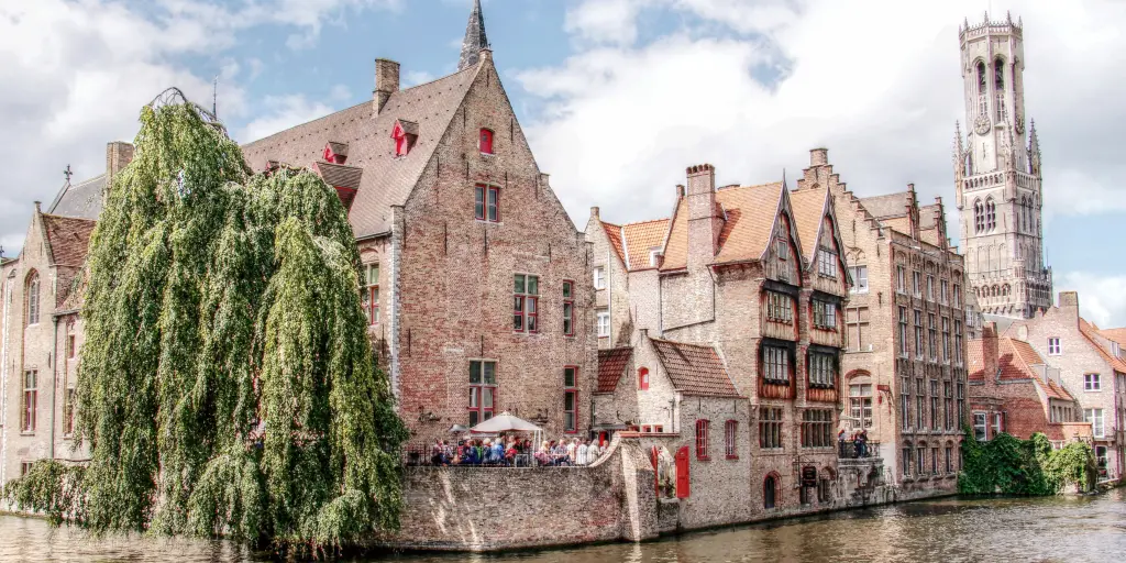 People stand under an umbrella at a restaurant on the river in Bruges, Belgium, with the belfry in the distance