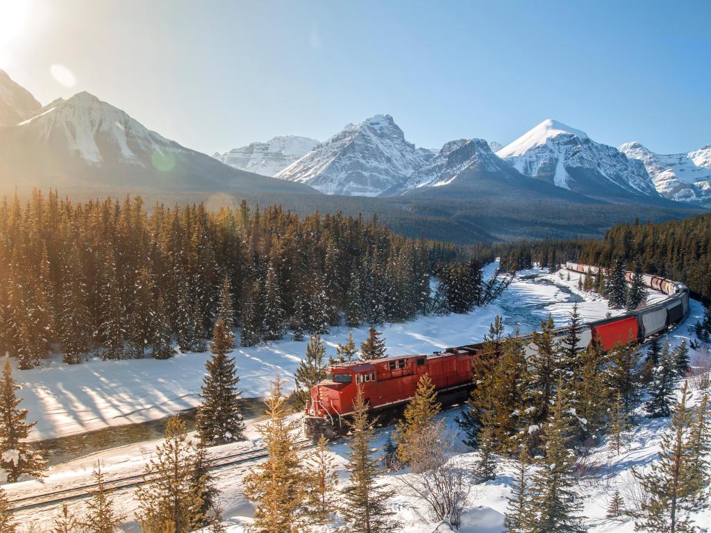 Red cargo train passing through snowy trees and mountains at Morant's curve near Banff, AB, Canada on a sunny winter's day
