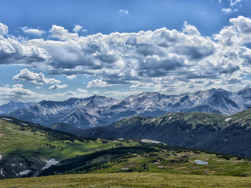 Rocky Mountain National Park, Colorado with rolling hills in the foreground and snow capped mountains in the background above a cloudy sky.