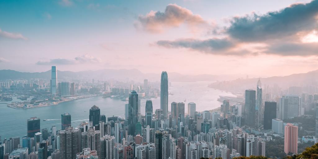 A view of the Hong Kong skyline and harbour from Victoria Peak, at sunset, with a pink and blue sky and high-rise buildings in the foreground