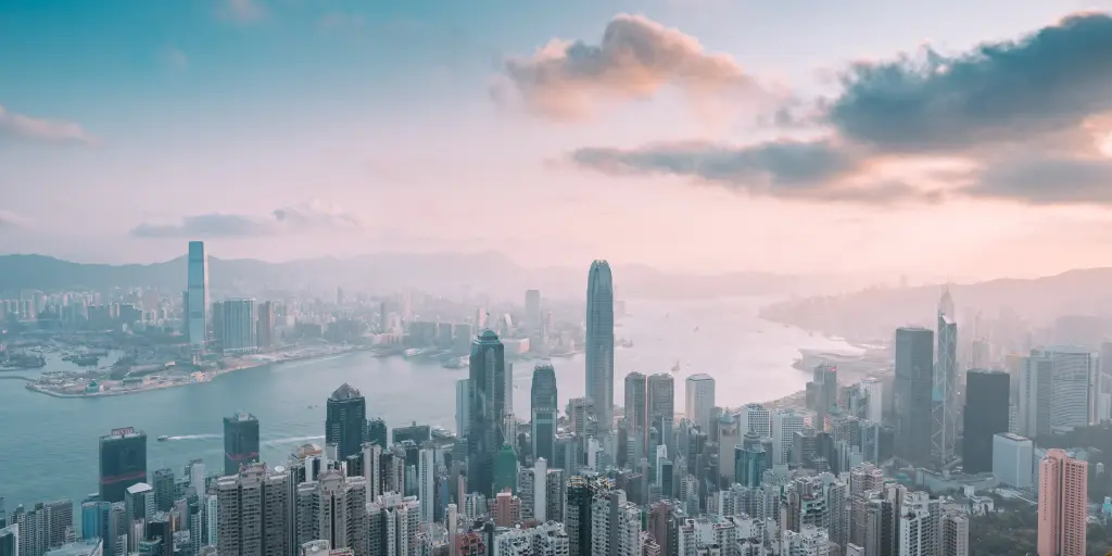 A view of the Hong Kong skyline and harbour from Victoria Peak, at sunset, with a pink and blue sky and high-rise buildings in the foreground