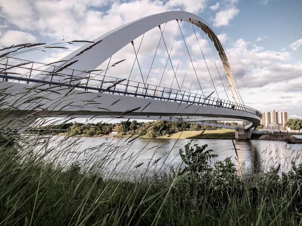 The photo depicts the Women of Achievement Bridge in Des Moines on a sunny day. There are some clouds in the sky and reeds are framing the corner of the photo.