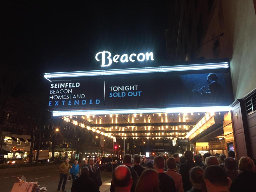 Exterior of The Beacon Theatre, with signs promoting Jerry Seinfeld performance and bright canopy lights. 