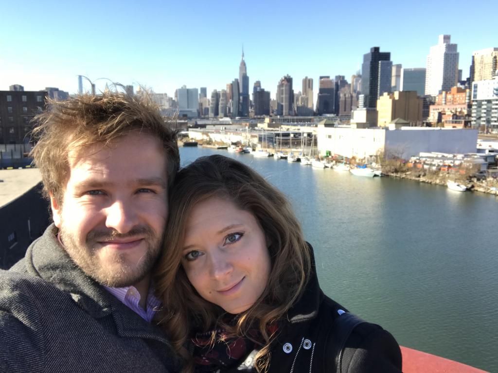 Sasha and Sarah standing on a bridge in Greenpoint, Brooklyn with the NYC skyline behind them.