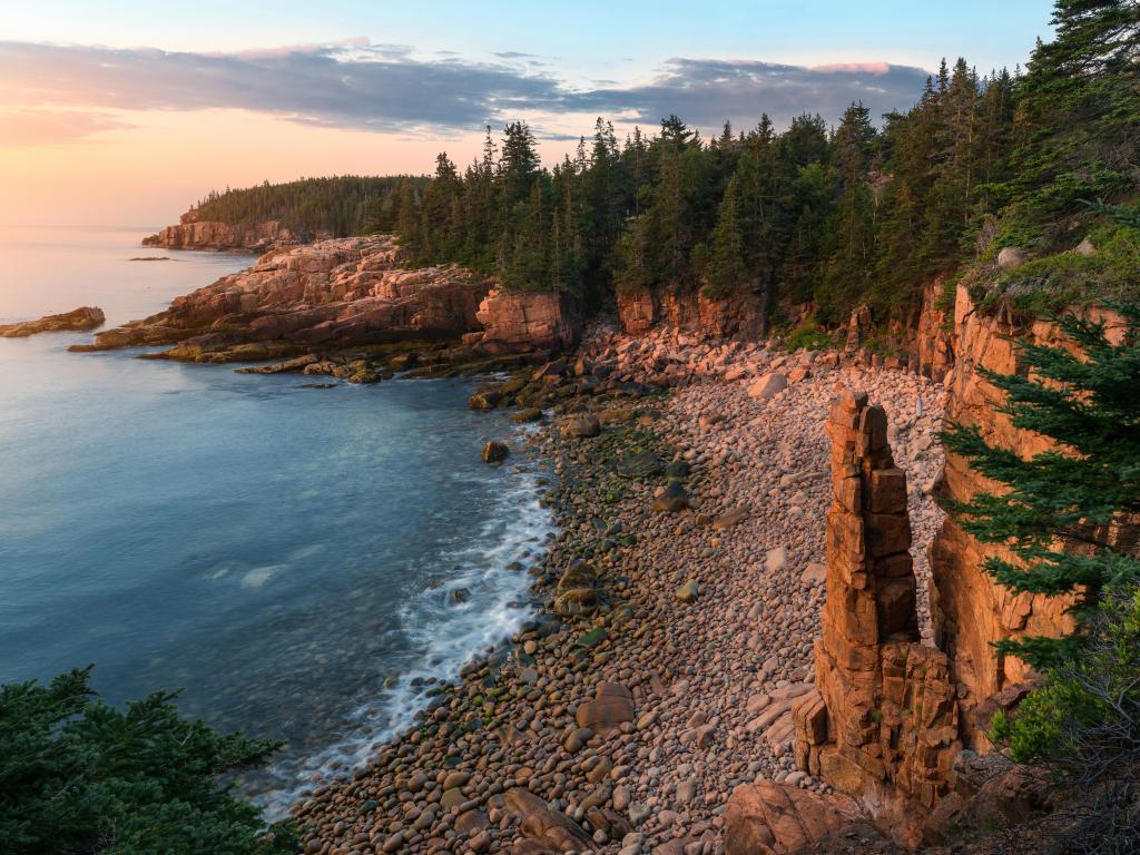 Acadia National Park, USA with the coast on the left and the rugged cliffs with tall trees above them on the right at sunrise.