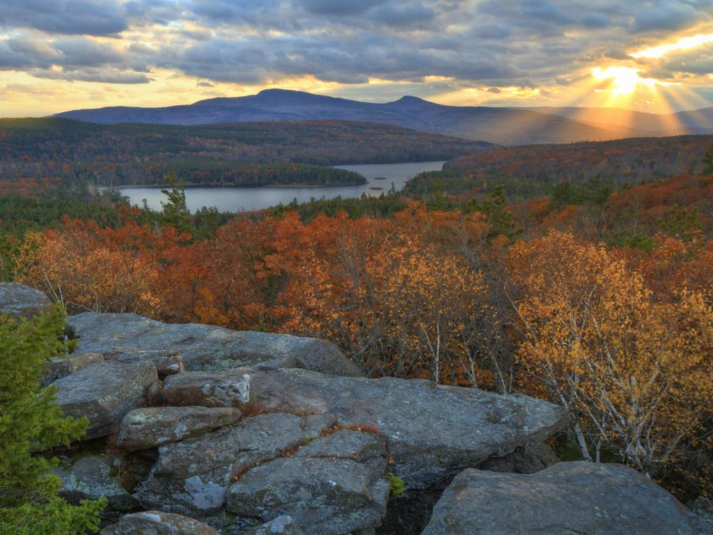Sunbeams escape the clouds at sunset across a valley covered in fall colored foliage, overlooking a Lake