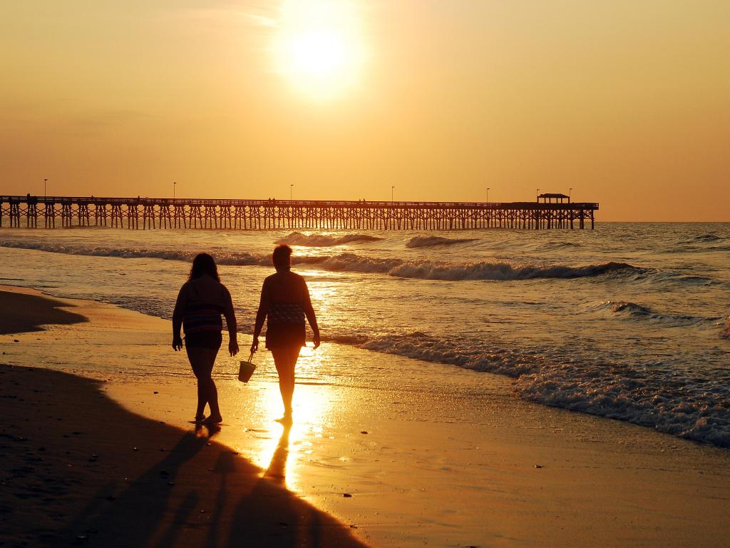 Two people strolling on the sandy shores of Myrtle Beach during a fiery, orange sunset.