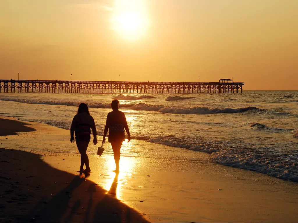 Two people strolling on the sandy shores of Myrtle Beach during a fiery, orange sunset.