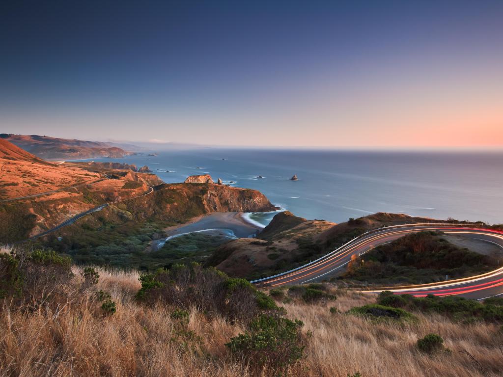 Ocean view with winding road travelling along cliffs through low late evening light with long exposure car lights shown on the road