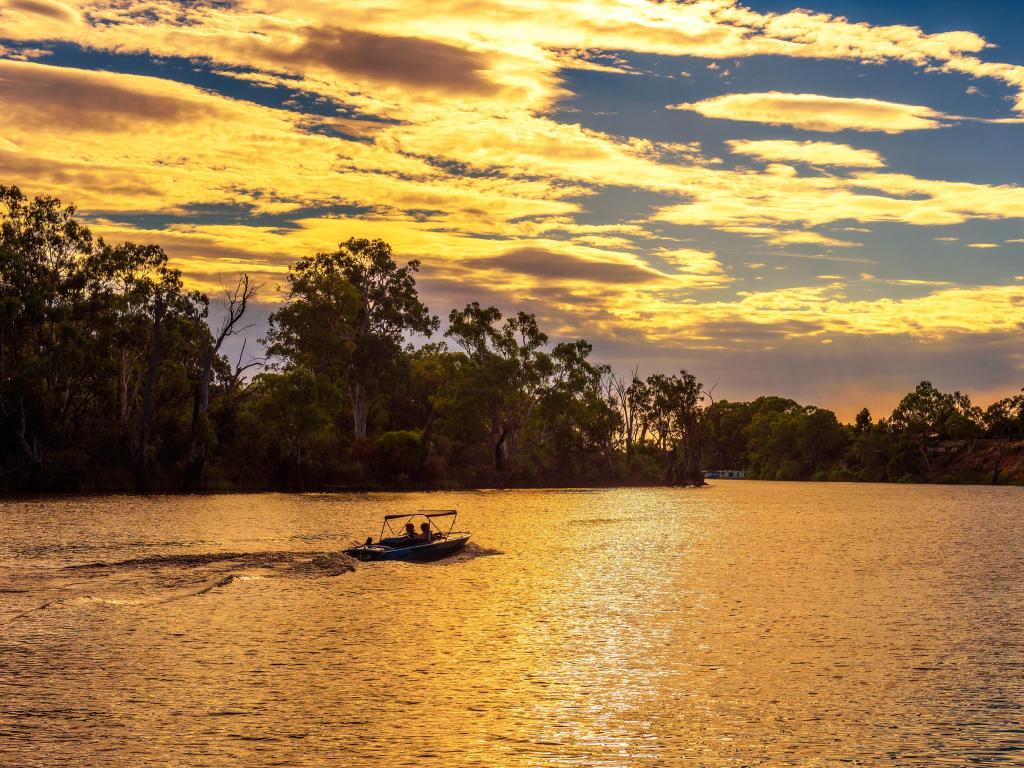 Wide river lit up in golden sunset light with silhouette trees on the bank and people riding in a small boat