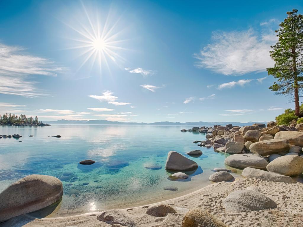 Lake Tahoe with a beach made of sand and large rocks to the left and a calm turquoise sea on a sunny day.