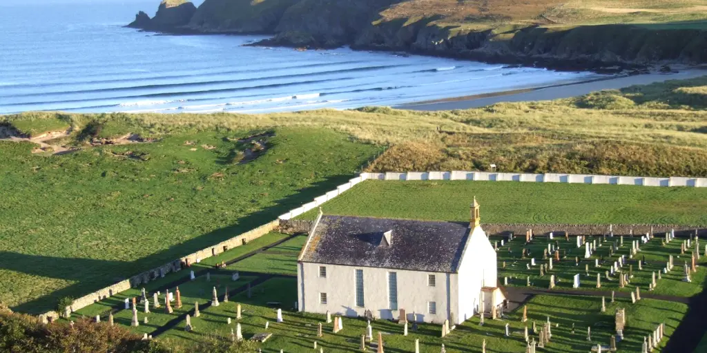 An aerial view of the white exterior of the Strathnaver Museum, Scotland, with a graveyard surrounding it, and a sandy cove and sea in the background