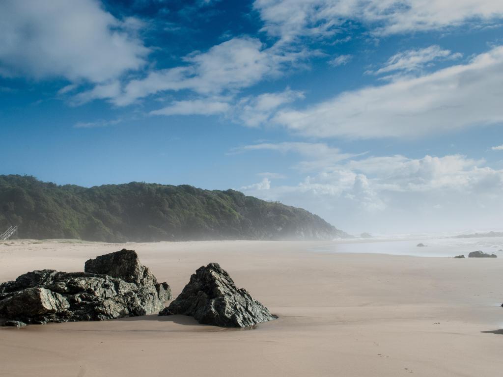 Wide golden sand beach with a single rock in the foreground and a headland covered in trees along the horizon