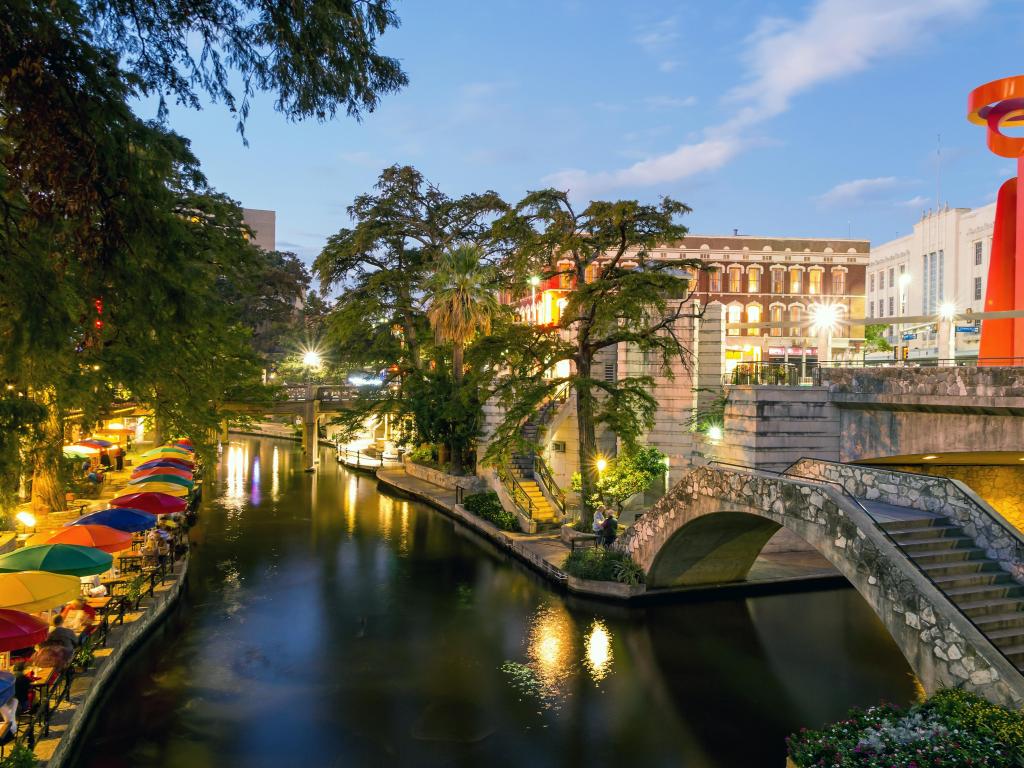 Colourful umbrellas and riverside seating with stone bridge and historic building lit up in evening light. Some trees.