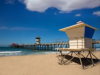 Huntington Beach Pier in background with lifeguard tower on sandy shore in foreground. Bright blue sky and wispy cloud overhead