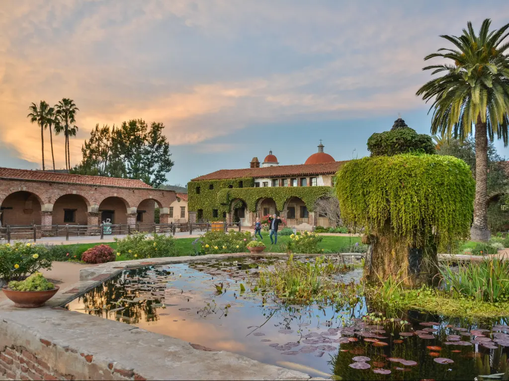 Courtyard of Mission San Juan Capistrano in California at dusk, with people and vegetation.
