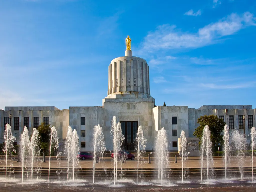 The Oregon State Capitol building in Salem with a fountain spouting water in front and a blue sky above.