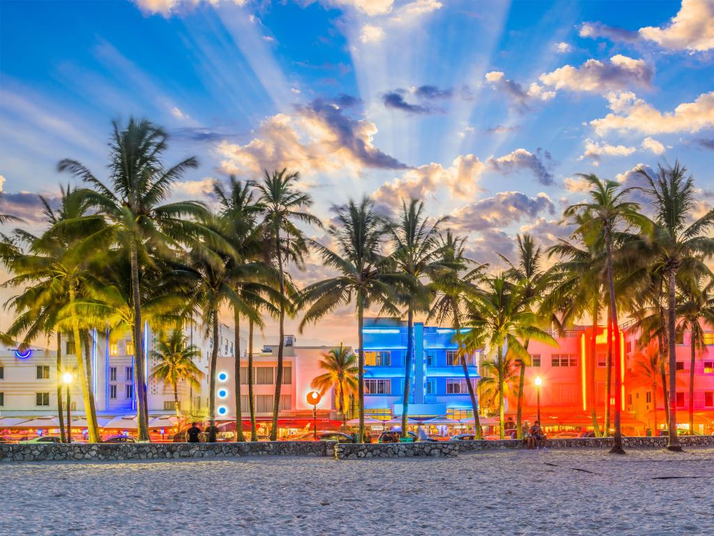 Beach with palm trees and brightly coloured buildings with rays of light from the setting sun behind them