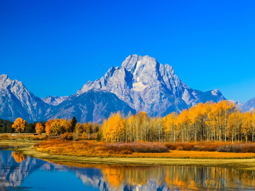 Clear blue sky, grey mountain with patches of snow, trees lit up orange in the sunlight, all reflected into still lake water