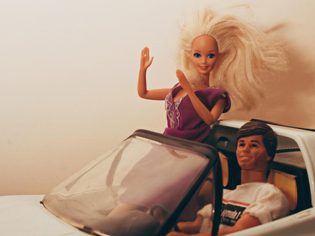 Barbie riding in a toy car with Ken, beige background