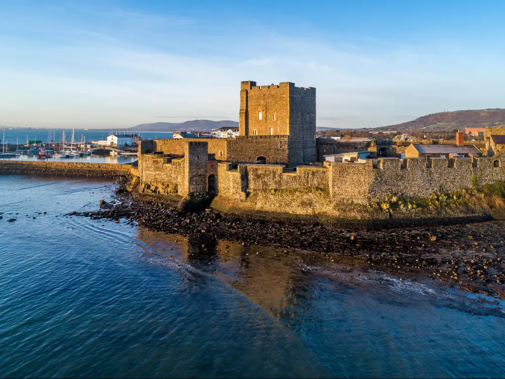 Medieval Norman Castle in Carrickfergus near Belfast in sunrise light. Aerial view with marina, yachts, breakwater, sediments and far view of Belfast in the background