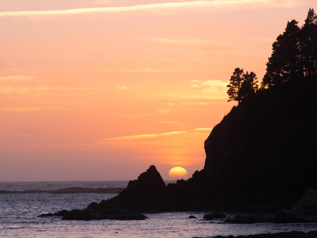 Sunset Bay State Park, Oregon with the sun setting behind a rocky point creating a silhouette and calm water in the foreground. 