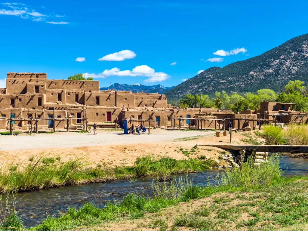 An image of a native pueblo in Taos, New Mexico. On sight is a building made of clay and on the right side is a portion of a mountain in a bright sunny day. A small river and some grasses are also in the image.