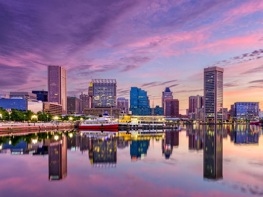 Baltimore, Maryland, USA with the city skyline in the distance reflecting in the water at the Inner Harbor in the foreground and taken after sunset with a purple sky.
