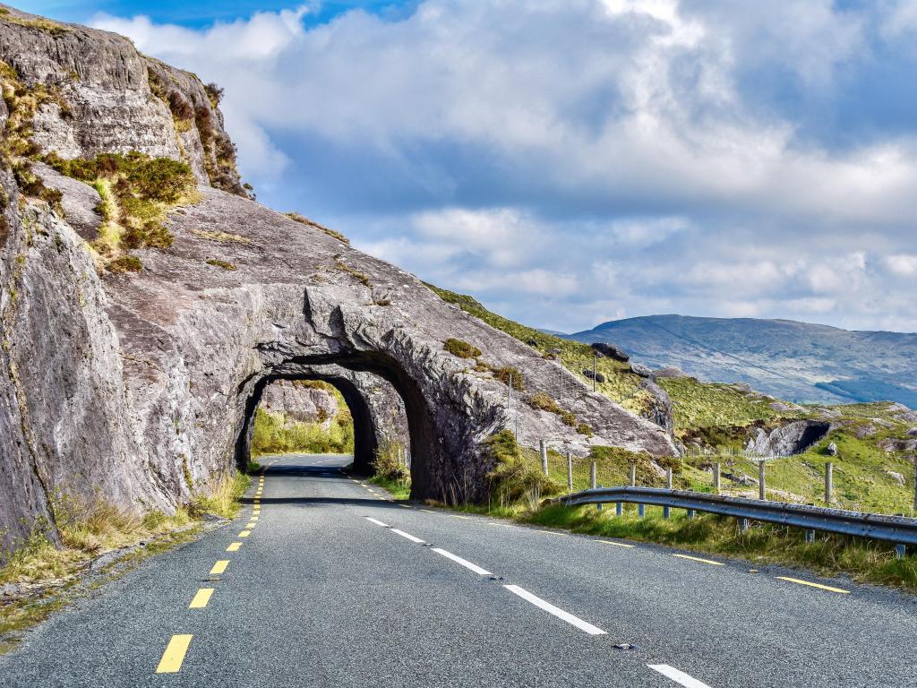 Kenmare Road, County Kerry, Ireland, stone arch tunnels made from gray stone showing lined highway N71 with gray guardrail in the foreground backed by light green mountains and a mostly cloudy sky.