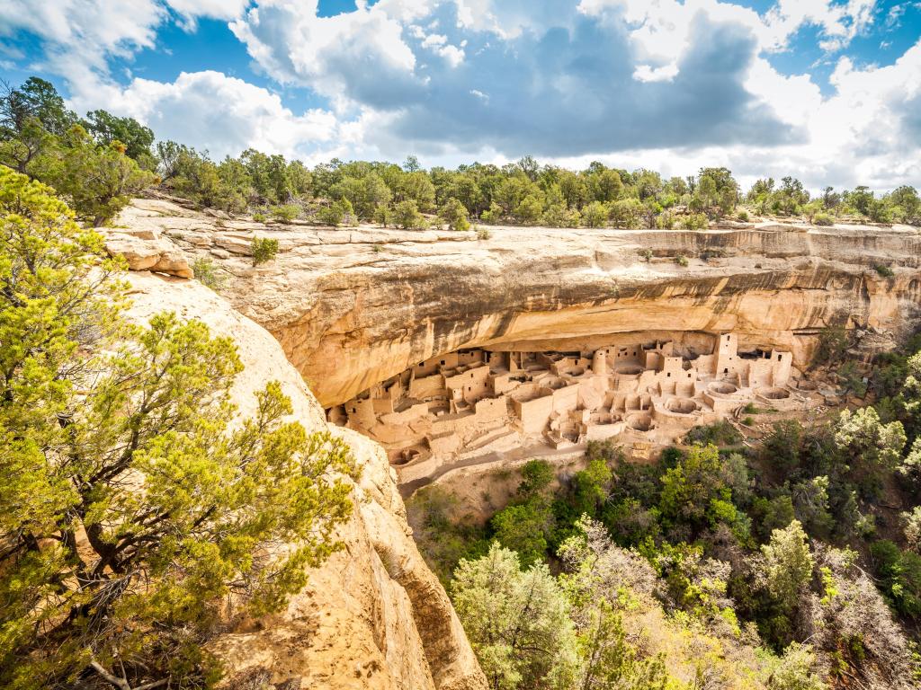 Mesa Verde National Parks, Colorado with cliffs overlooking a valley covered in trees below and over a cloudy blue sky.