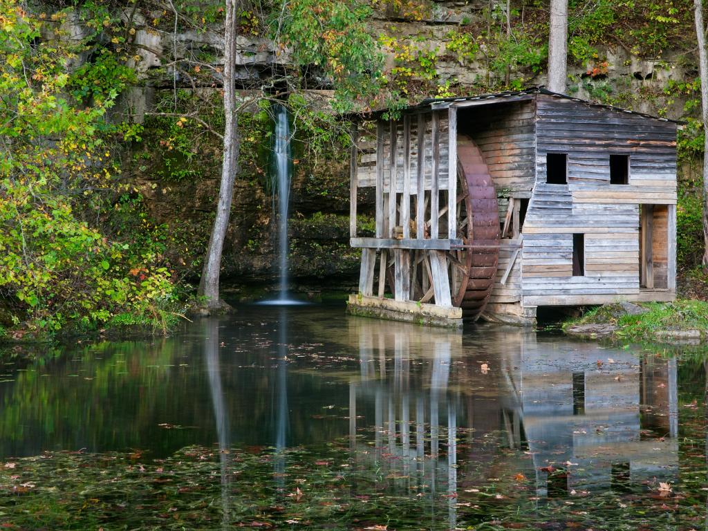 Old wooden water mill with green vegetation and a small waterfall, reflected into the still lake